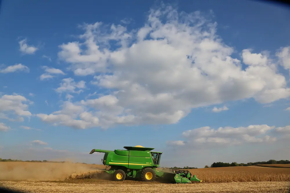 Green combine harvester harvesting grain in a field with dust clouds under a partly cloudy sky